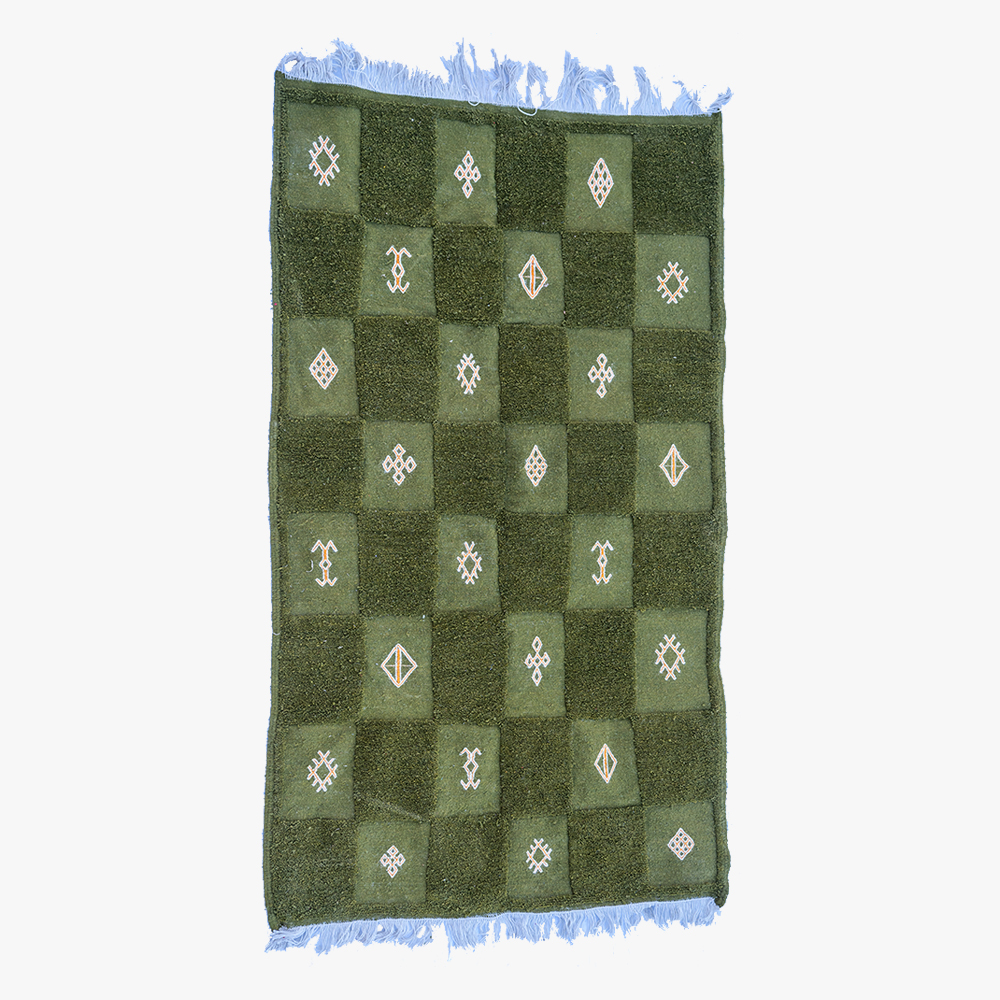 Groen berber tapijt met schaakbordpatroon en witte geometrische symbolen, omzoomd met lichtblauwe franjes.